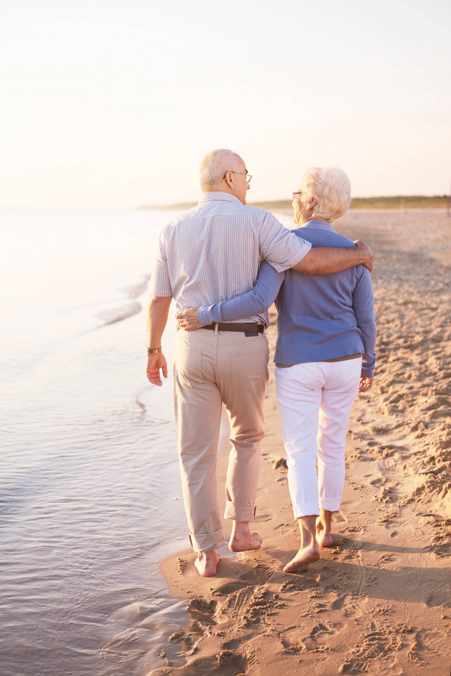 two-senior-adults-by-ocean_11zon Helbredstjek 70 år: Portræt af en mand i hans 70'ere, der står iført en hvid T-shirt og sorte bukser med seler. Han smiler mod kameraet.